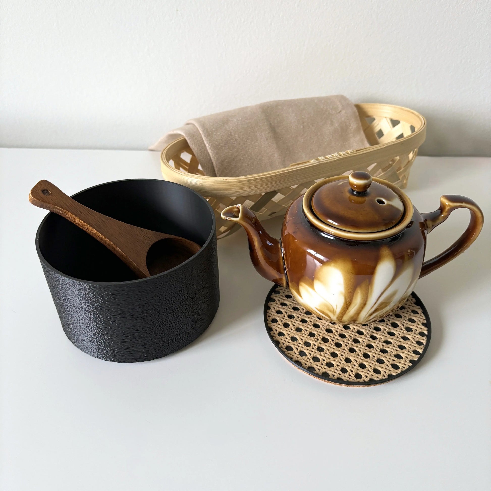 Black COZI minimalist canister on a kitchen counter, holding a teapot and cup on its rattan lid by Sonnihaus